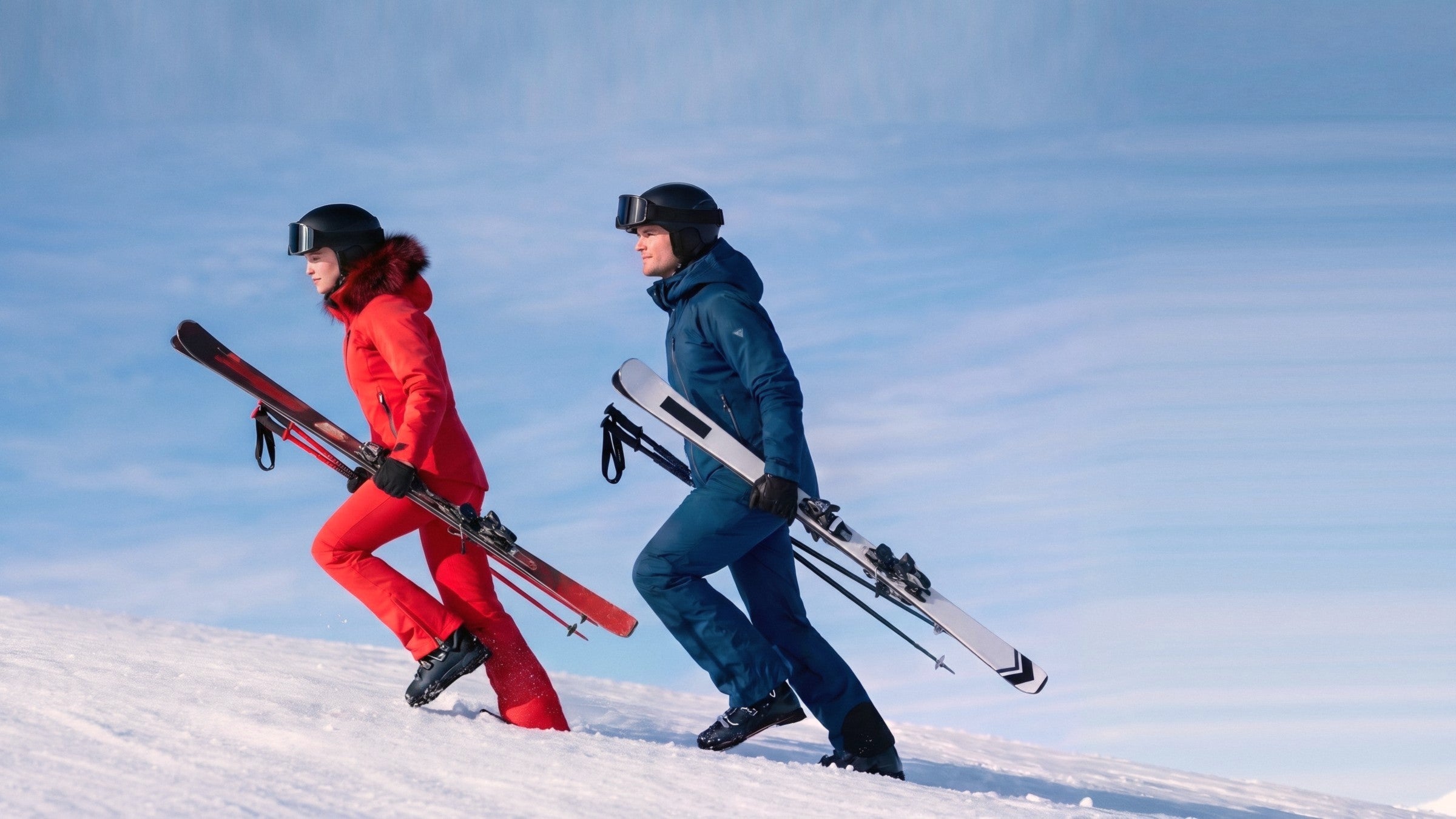 Pareja de esquiadores con chaquetas Tsunami roja y azul marino subiendo con los esquís por la nieve