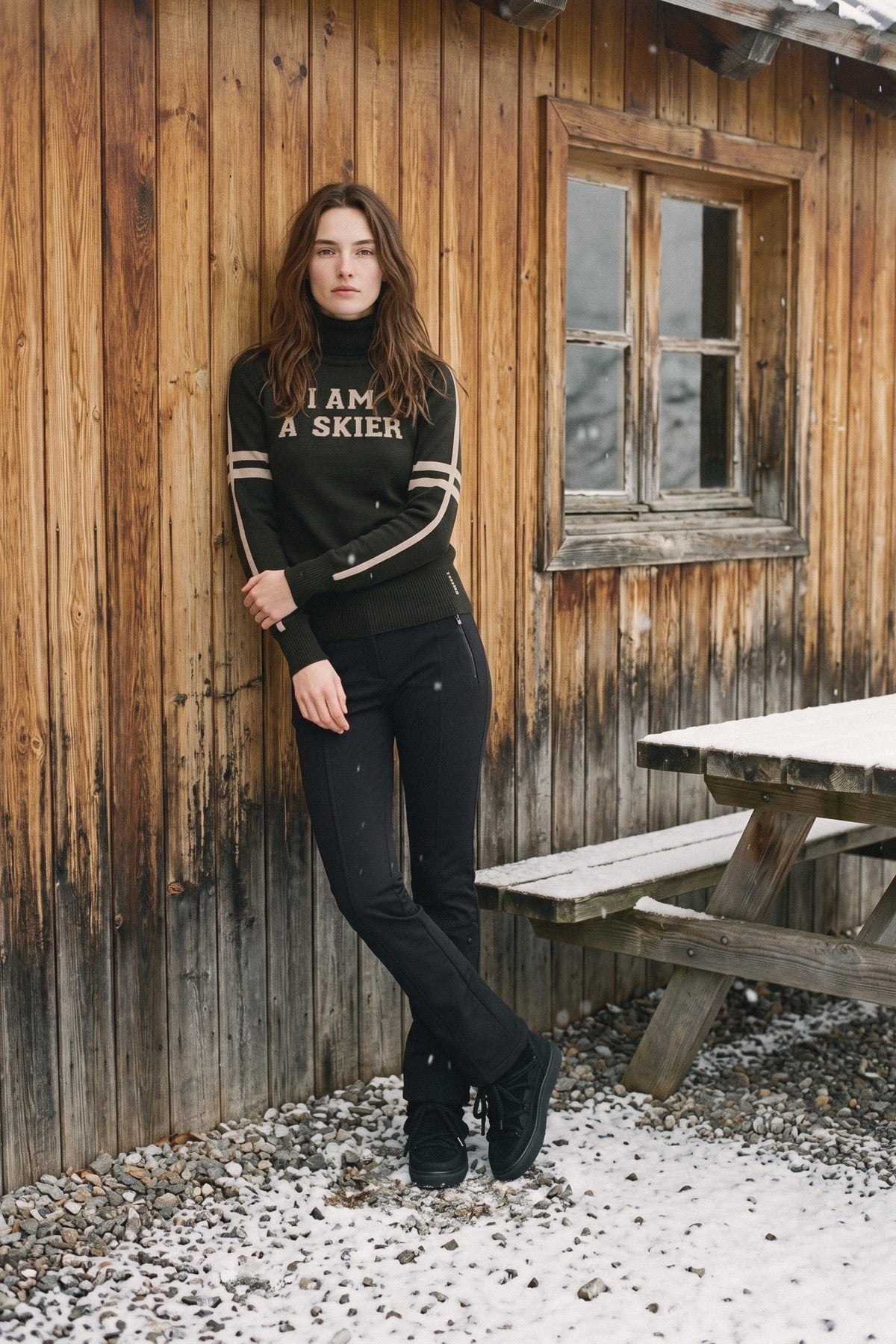 Mujer con el jersey Tsunami de lana merino negro «I AM A SKIER» junto a una cabaña de madera nevada